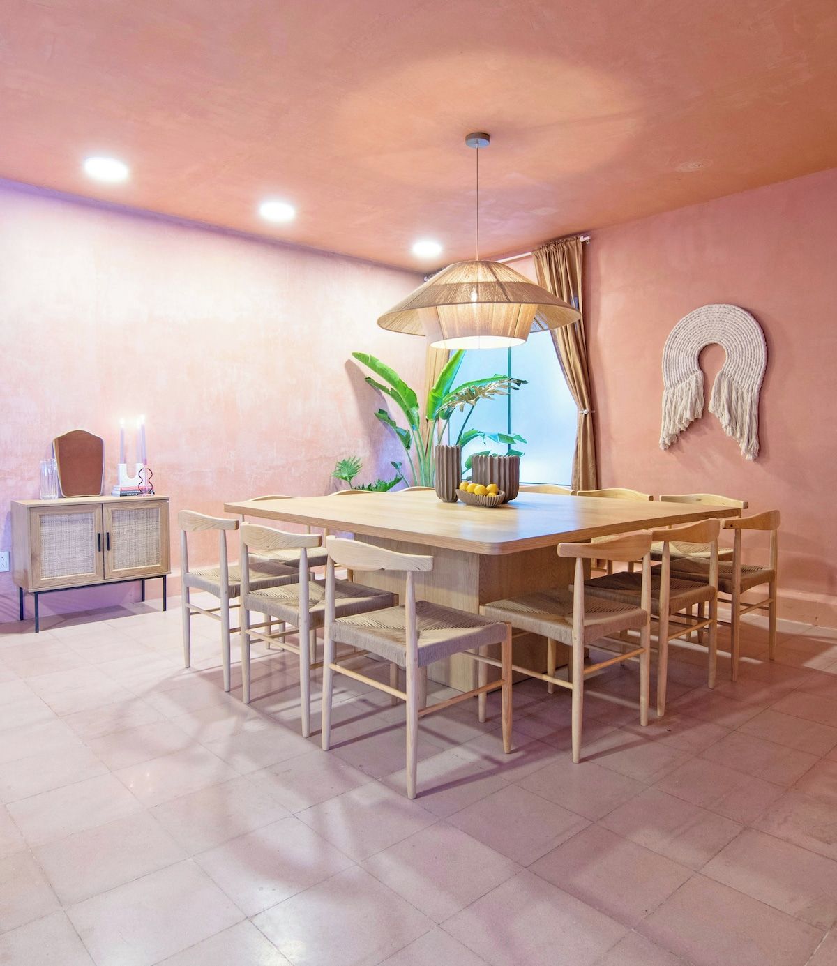 A dining area with a wooden table and chairs, a hanging lamp, and a macrame wall decoration.