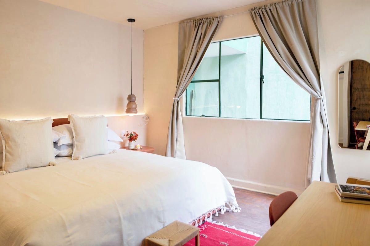 Quarto com cama grande, mesa de cabeceira com abajur, janela com cortinas e pequena mesa com cadeira.