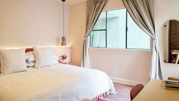 Quarto com cama grande, mesa de cabeceira com abajur, janela com cortinas e pequena mesa com cadeira.