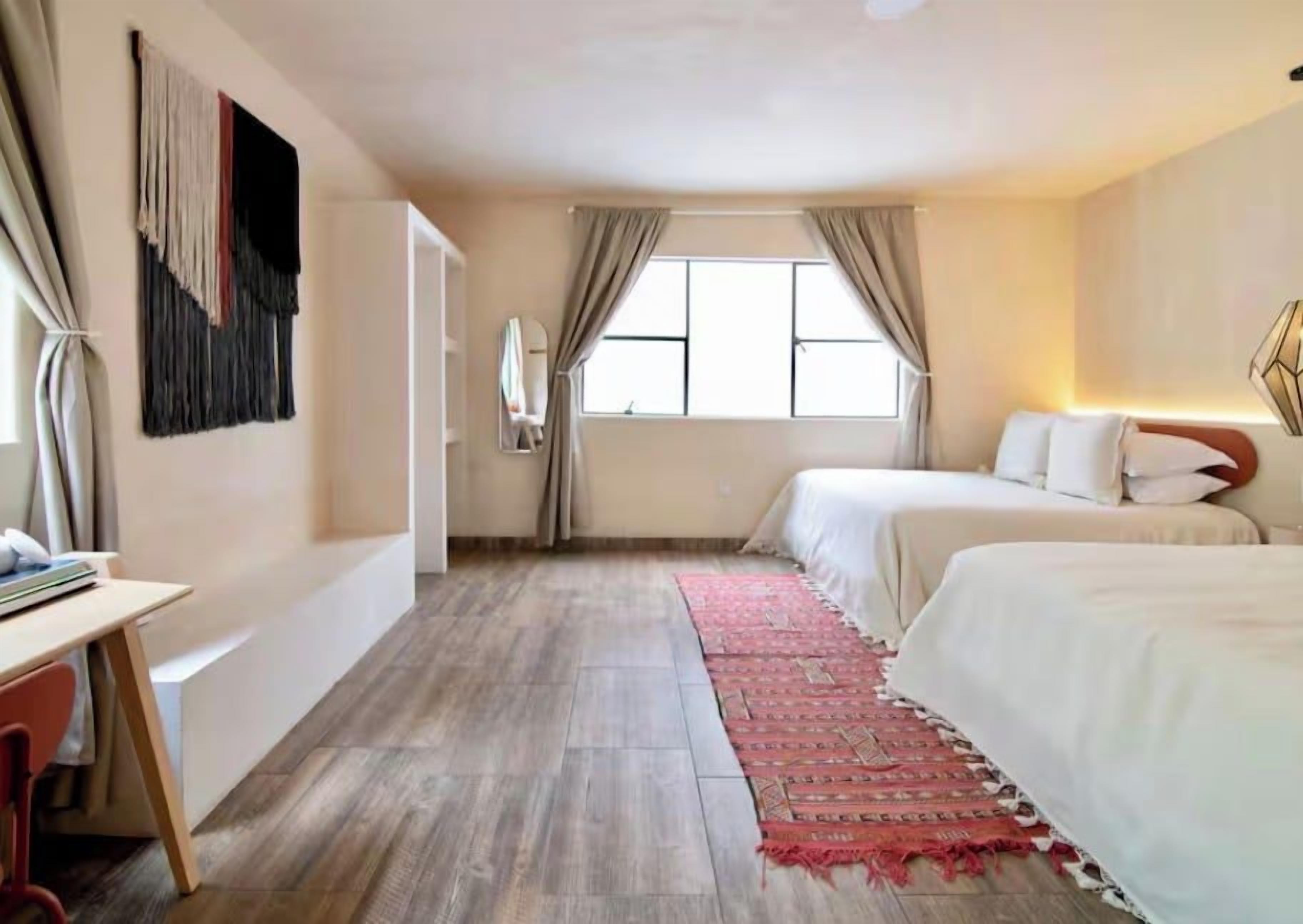 Ein Hotelzimmer mit Bett, Schreibtisch, Stuhl, einem Fenster mit Vorhängen und einem an der Wand hängenden Kunstwerk.