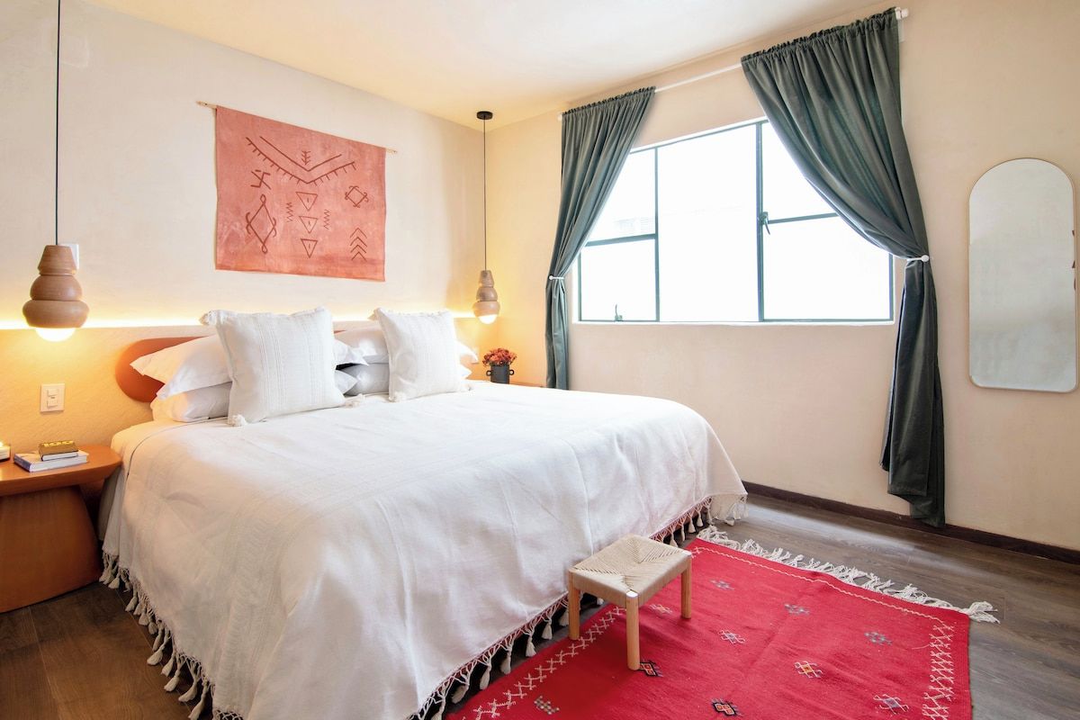 A hotel room with a bed, bedside table, lamp, window with curtains, and a wall tapestry.