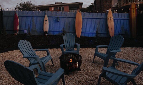 A backyard with a fire pit, three Adirondack chairs, and surfboards on a blue fence.