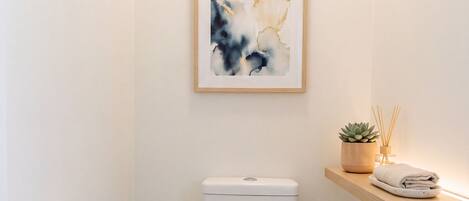 Een moderne badkamer met een toilet, een lijst met een abstract schilderij en een klein plankje met een plant en een handdoek.