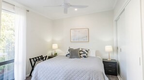 Un dormitorio con cama, dos mesitas de noche con lámparas, un ventilador de techo, una ventana con cortinas e imagen enmarcada en la pared.