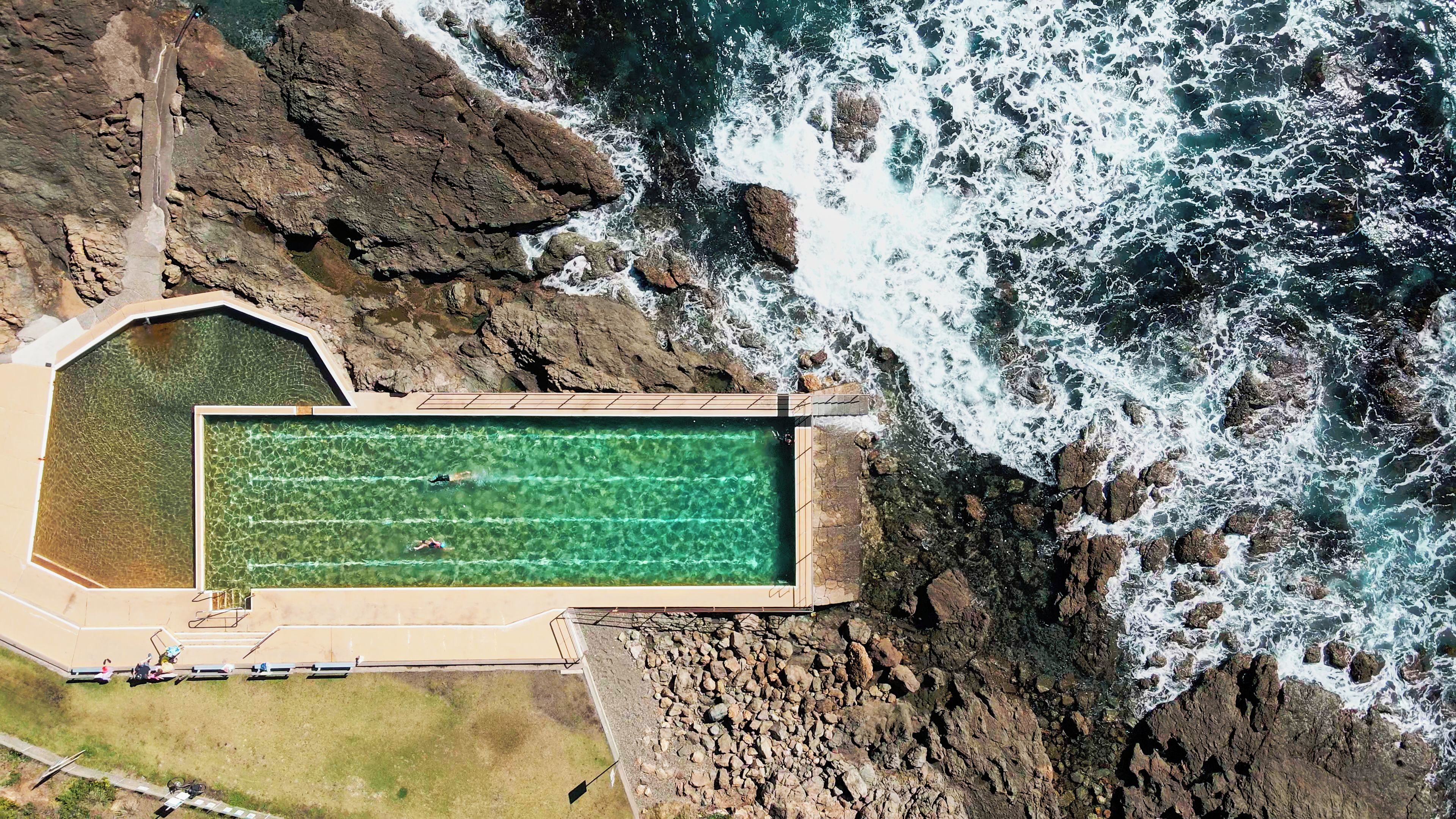 An aerial view of a swimming pool adjacent to a rocky coastline with waves crashing onto the shore.