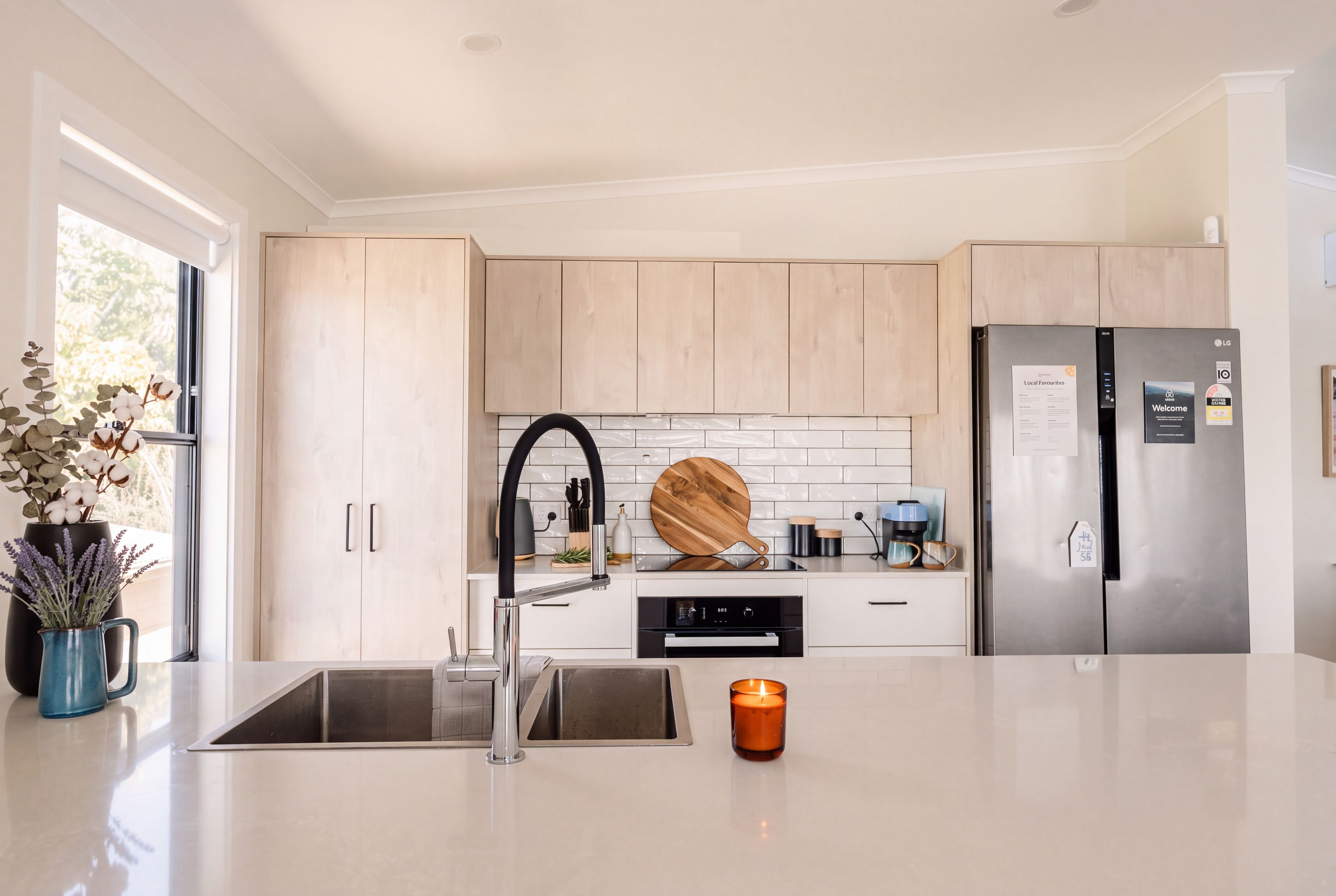 A modern kitchen with light wood cabinets, a stainless steel refrigerator, a built-in oven, and a large island with a sink.