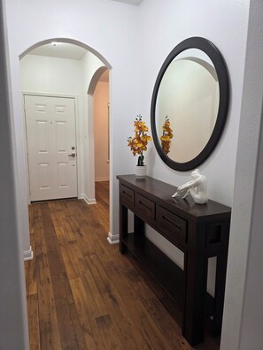 A hallway with a wooden floor, a white door, a dark wooden console table with a mirror, and a vase with flowers. - Charming 3-bedroom house with AC in Rosharon (Rosharon)