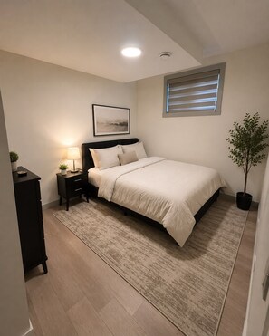 A bedroom with a bed, bedside tables, a lamp, a picture on the wall, a window with blinds, and a potted plant. - The Bowness Haven (Calgary)