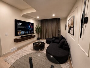 A modern living room with a large flat-screen TV, a black sectional sofa, a round coffee table, and a potted plant. - The Bowness Haven (Calgary)