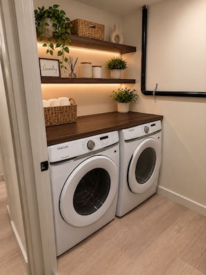 A laundry room with two Samsung washing machines, wooden shelves with plants and baskets, and a wall-mounted pipe.