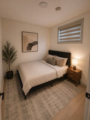 A bedroom with a bed, bedside table, lamp, potted plant, and a wall-mounted abstract painting. - The Bowness Haven (Calgary)