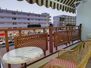Un balcon avec une table ronde et des chaises en osier, donnant sur une rue bordée d’immeubles et de boutiques.