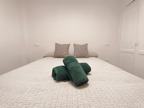 Een enkel bed met een wit geweven beddekleed en twee kussens, een groene handdoek en een witte garderobe met deur.