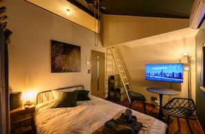 Habitación de hotel con cama, escritorio con lámpara, televisor mostrando 'Fukuoka City', una escalera y una silla.
