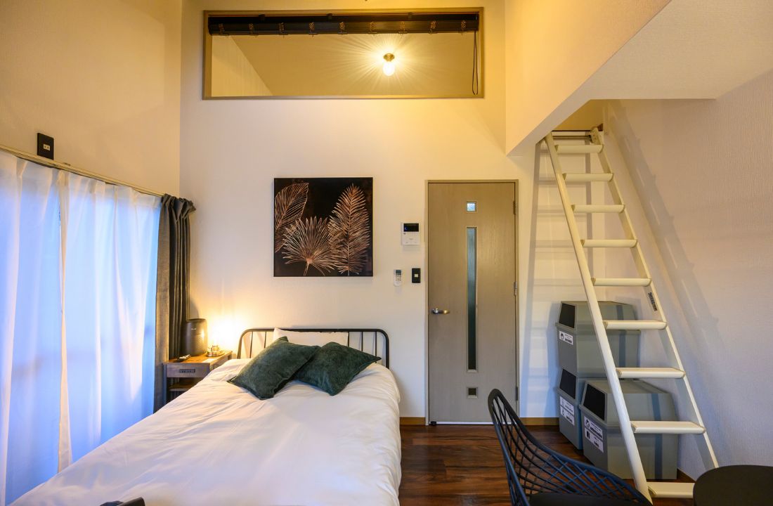 A bedroom with a bed, a nightstand, a wall-mounted light, a door, and a white ladder.
