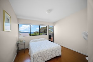 A bedroom with a bed, a nightstand, a lamp, a window with a view of the sea, and a ceiling fan. - Headland Home, modern 3 B/r perched on Bennetts Headland looking north & south. (Forster)