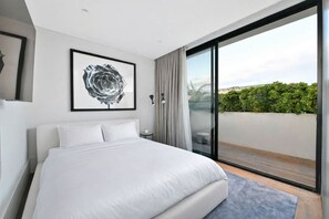 Ein modernes Hotelzimmer mit einem großen Bett, einem gerahmten Kunstwerk, Blick ins Freie und einer Schiebeglastür.