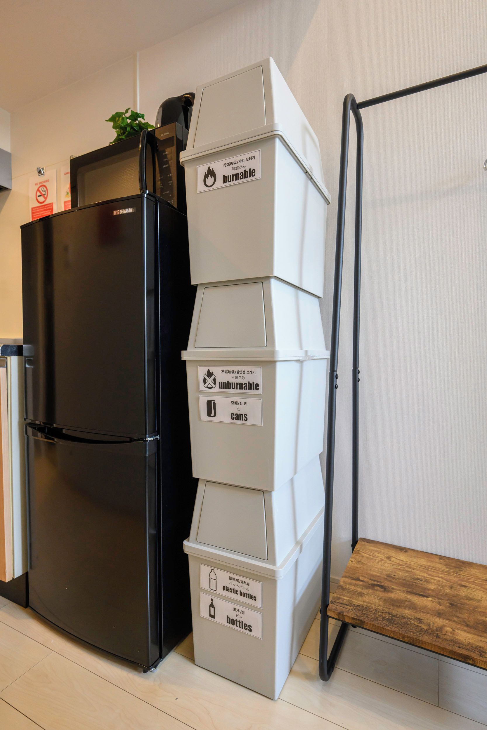 A black refrigerator next to a stack of white bins labeled for recycling, with a metal rack beside them.