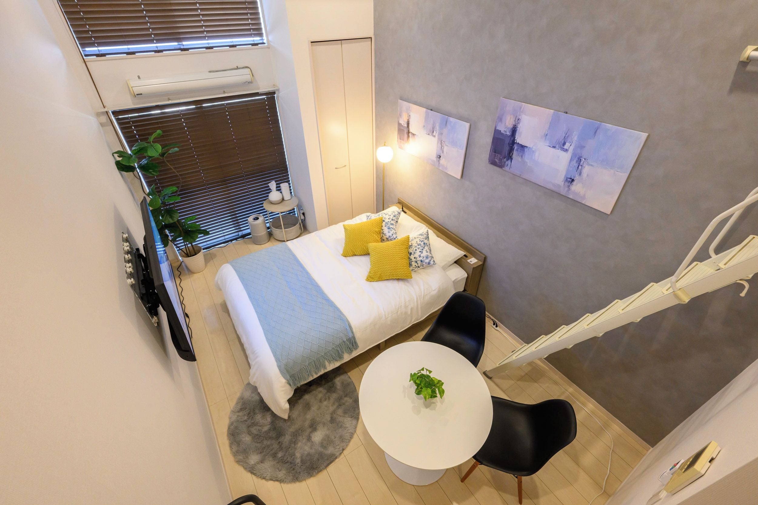 A hotel room with a bed, bedside table, lamp, two paintings, a round table, a chair, a potted plant, and a window with blinds.