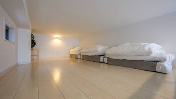 Una habitación con techo inclinado, una cama con ropa blanca, un taburete de madera pequeño y una lámpara fijada a la pared.