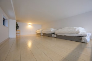 Una habitación con techo inclinado, una cama con ropa blanca, un taburete de madera pequeño y una lámpara fijada a la pared.