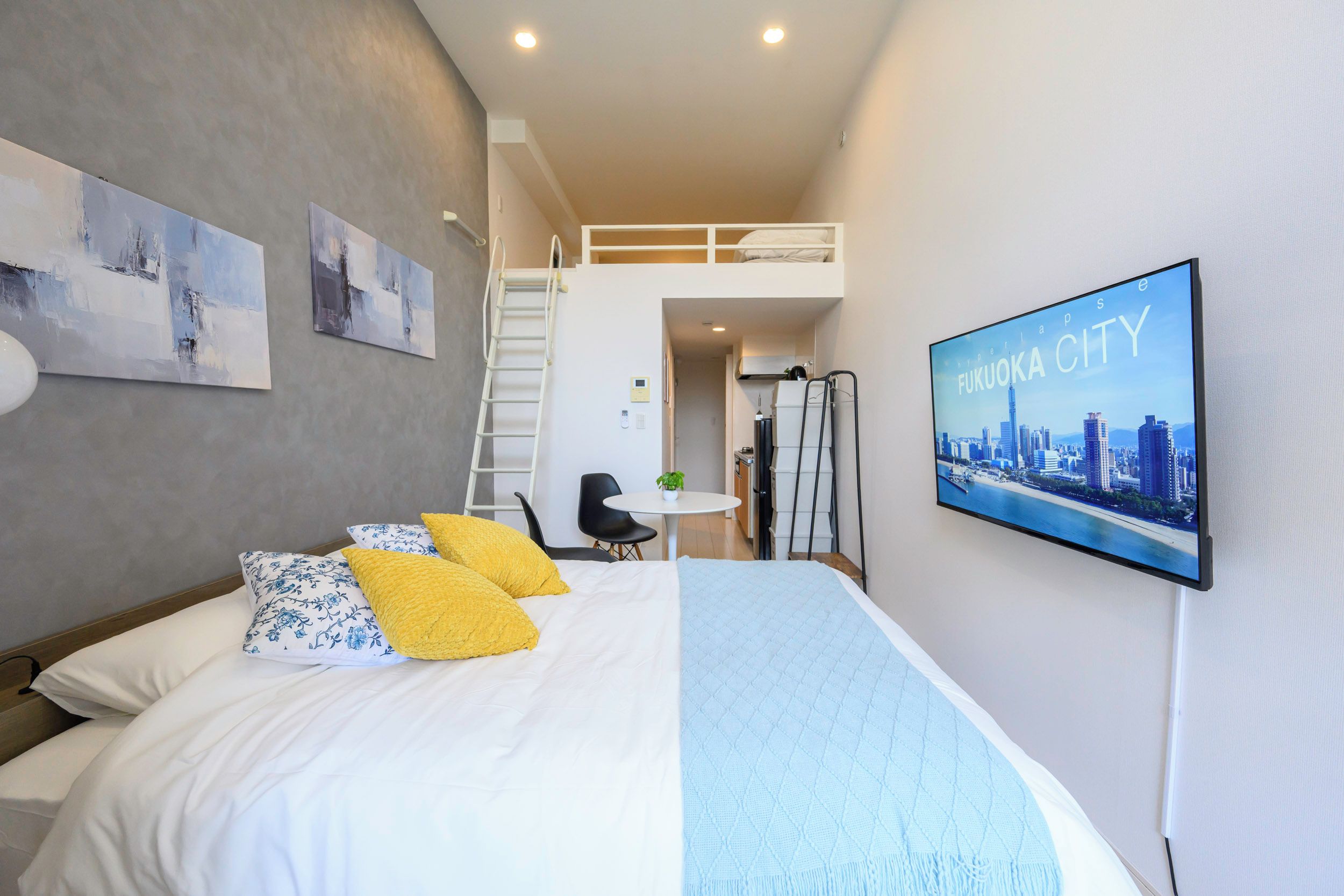 A hotel room with a bed, a desk, a chair, a ladder, a TV, and a cityscape picture on the wall.