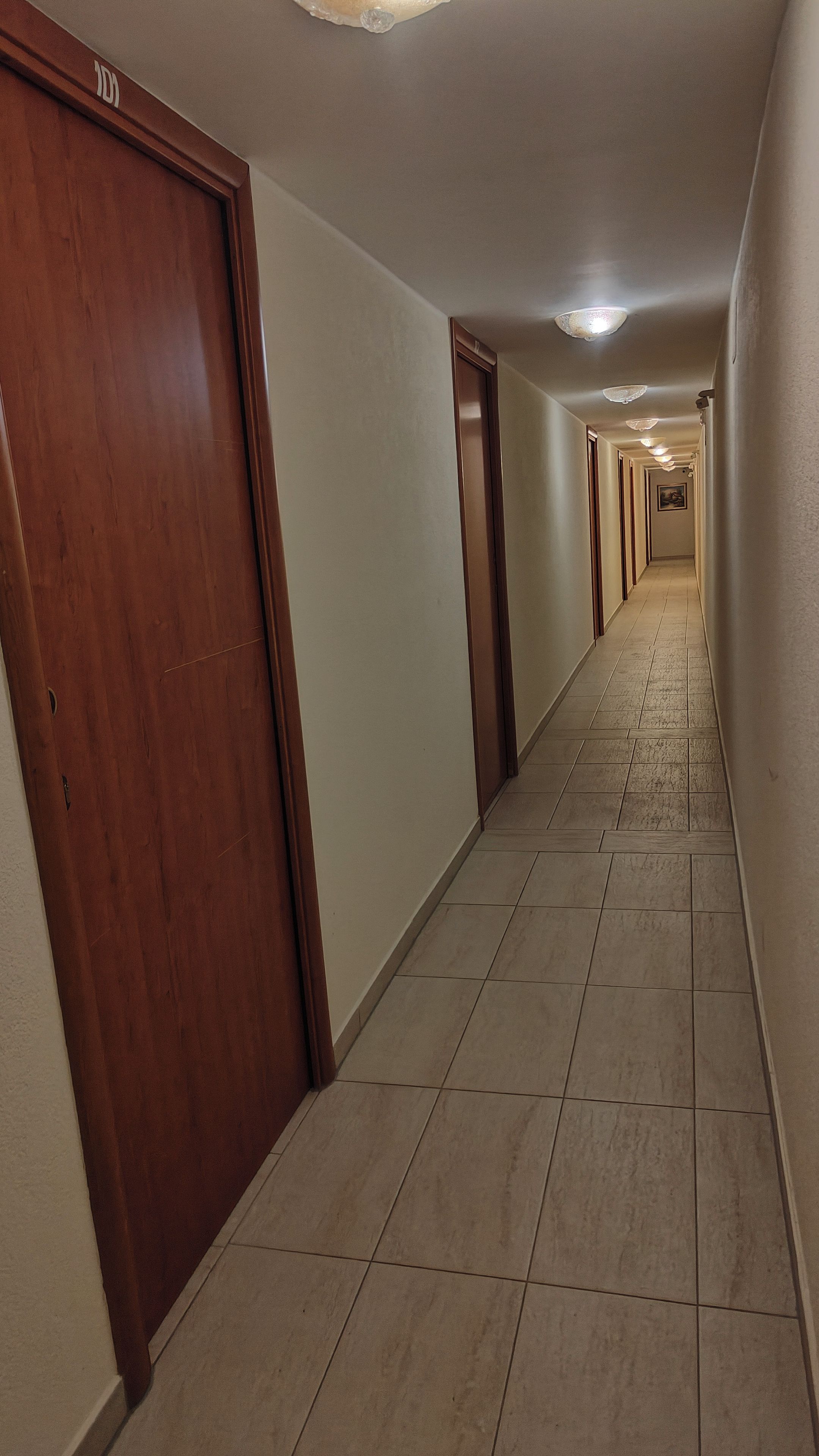 A corridor with wooden doors and tiled flooring, leading to a hallway with a ceiling light fixture.