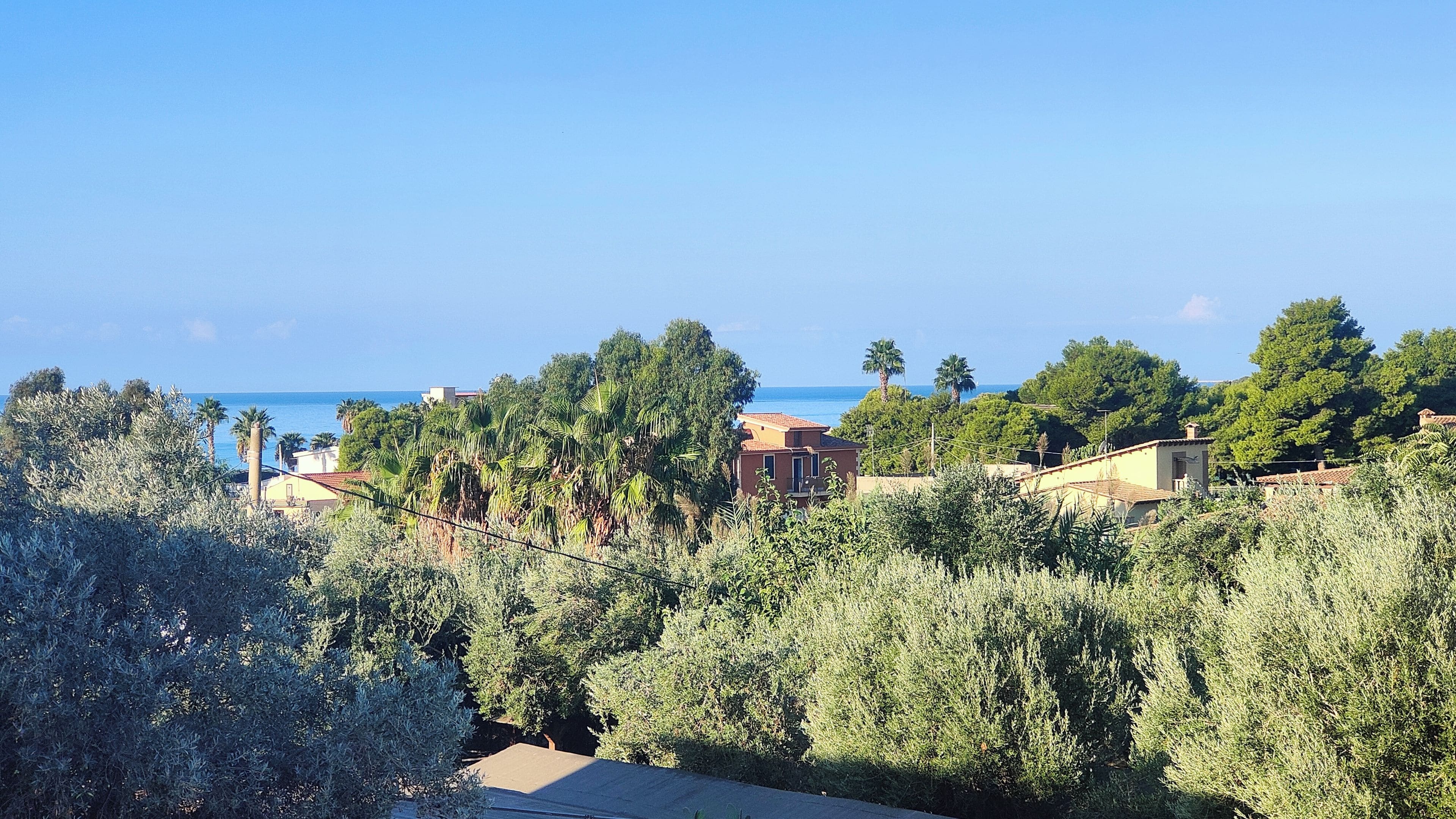 A coastal residential area with houses, dense greenery, and a clear view of the sea.
