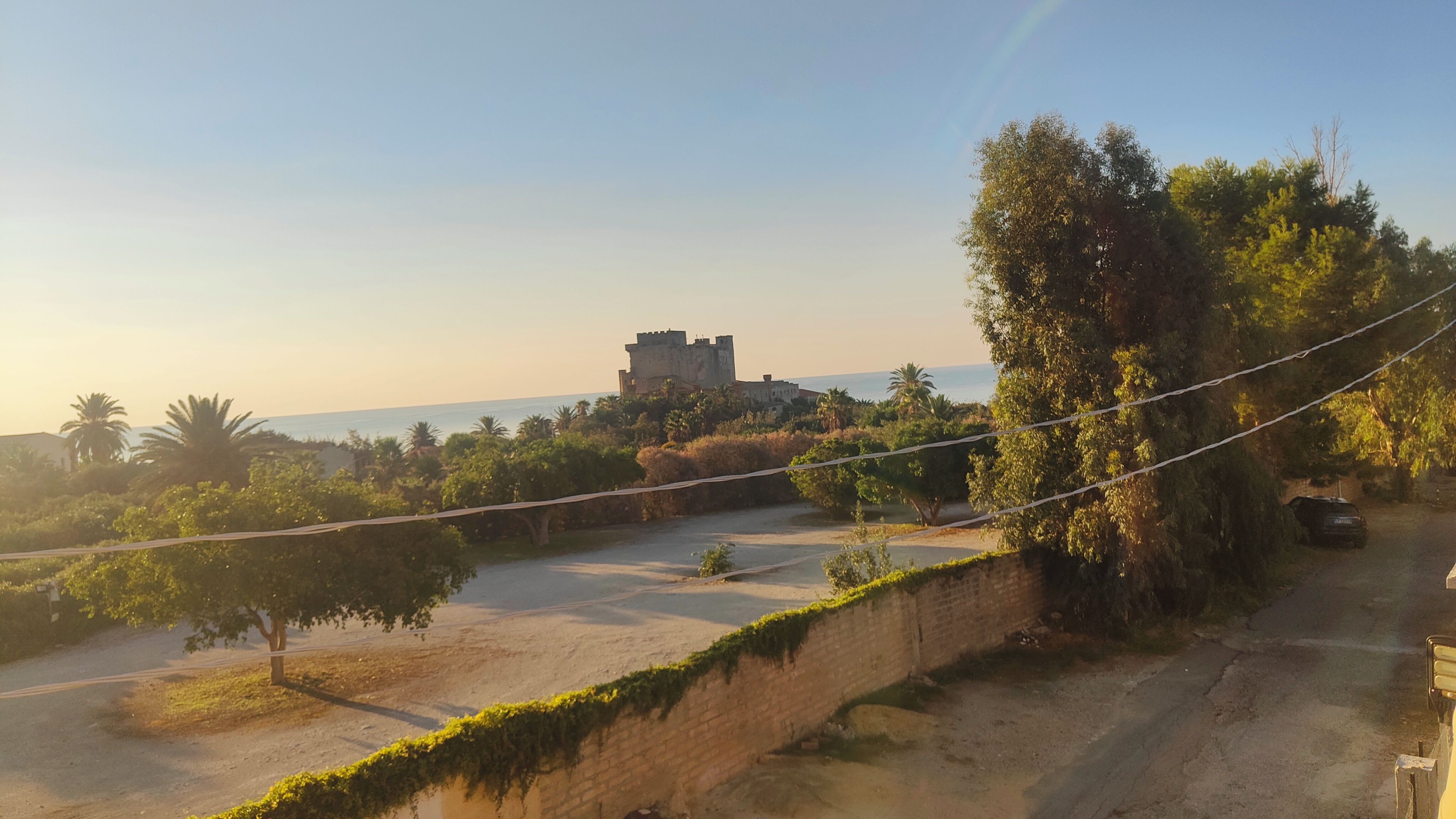 Un castillo en una colina con vistas despejadas al mar, rodeado de vegetación y un camino pavimentado.