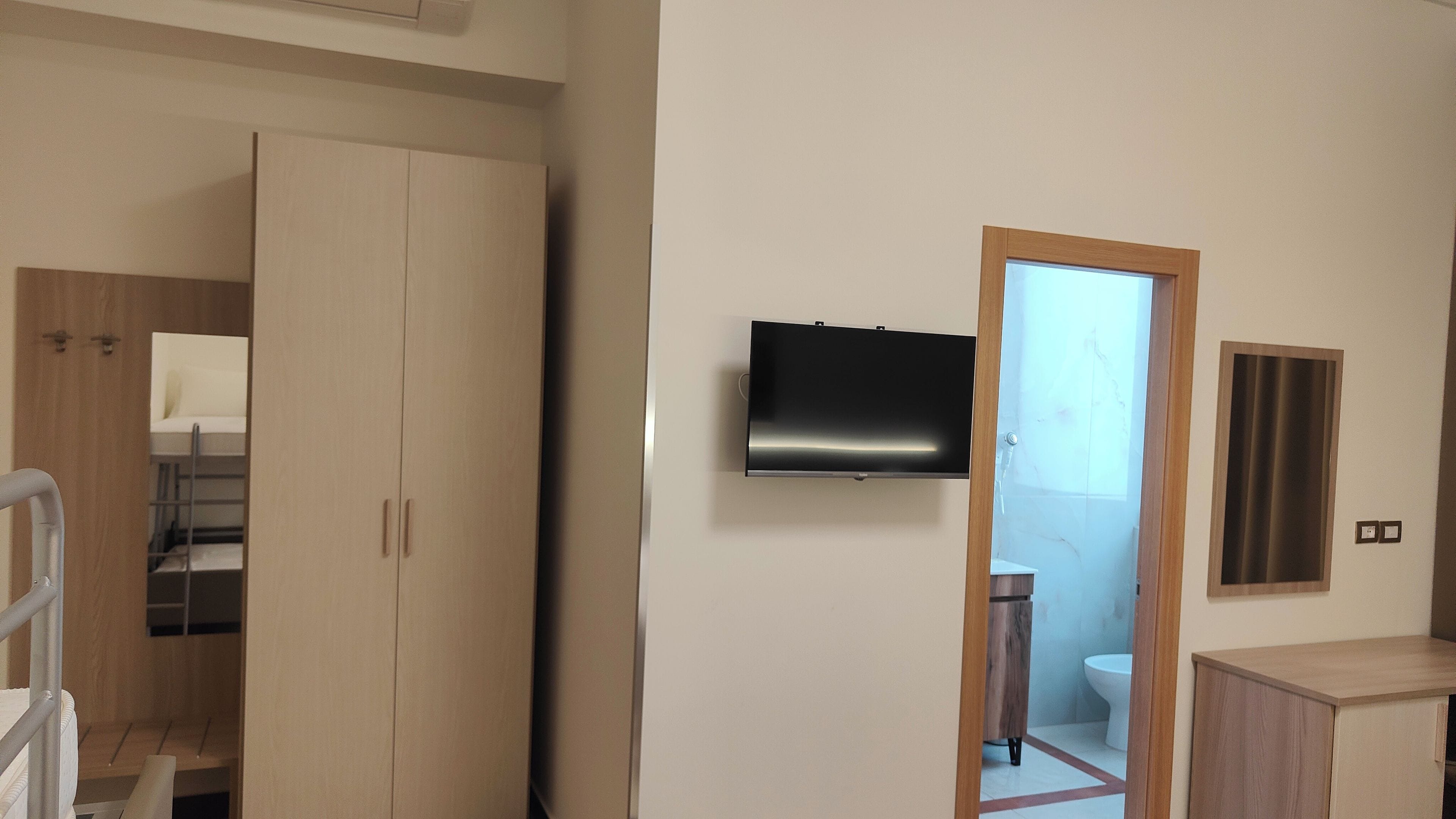 Ein Zimmer mit einem Kleiderschrank, einem an der Wand montierten Fernseher und einem durchgegangenen Tür sichtbaren Badezimmer.