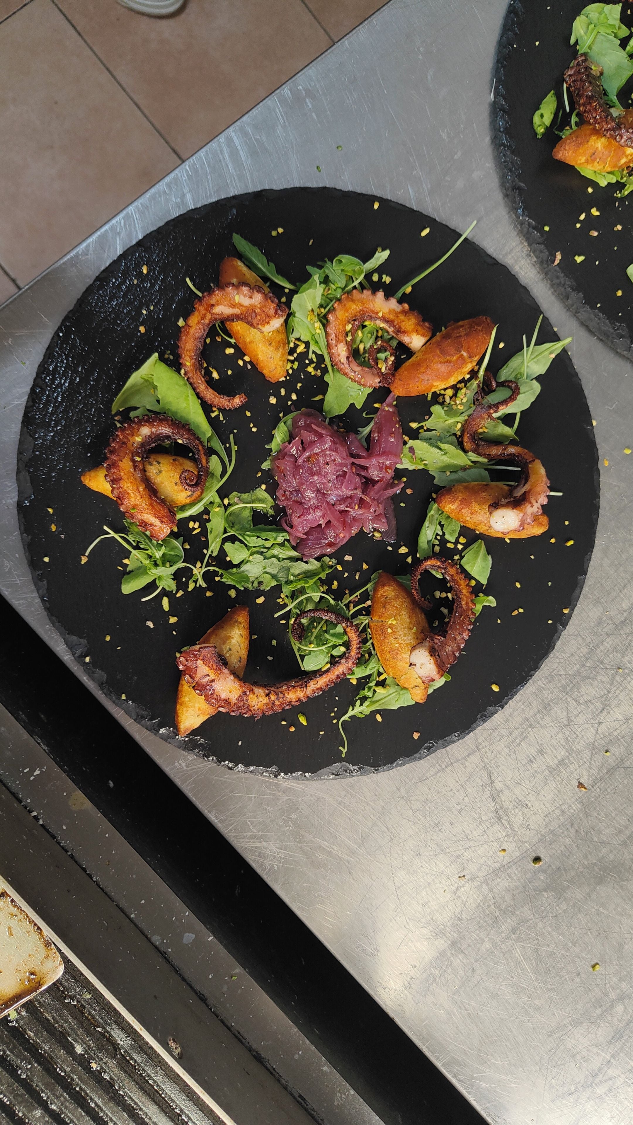 Un plato de pizarra negra con comida dispuesta en un patrón circular, rodeado por una superficie metálica.