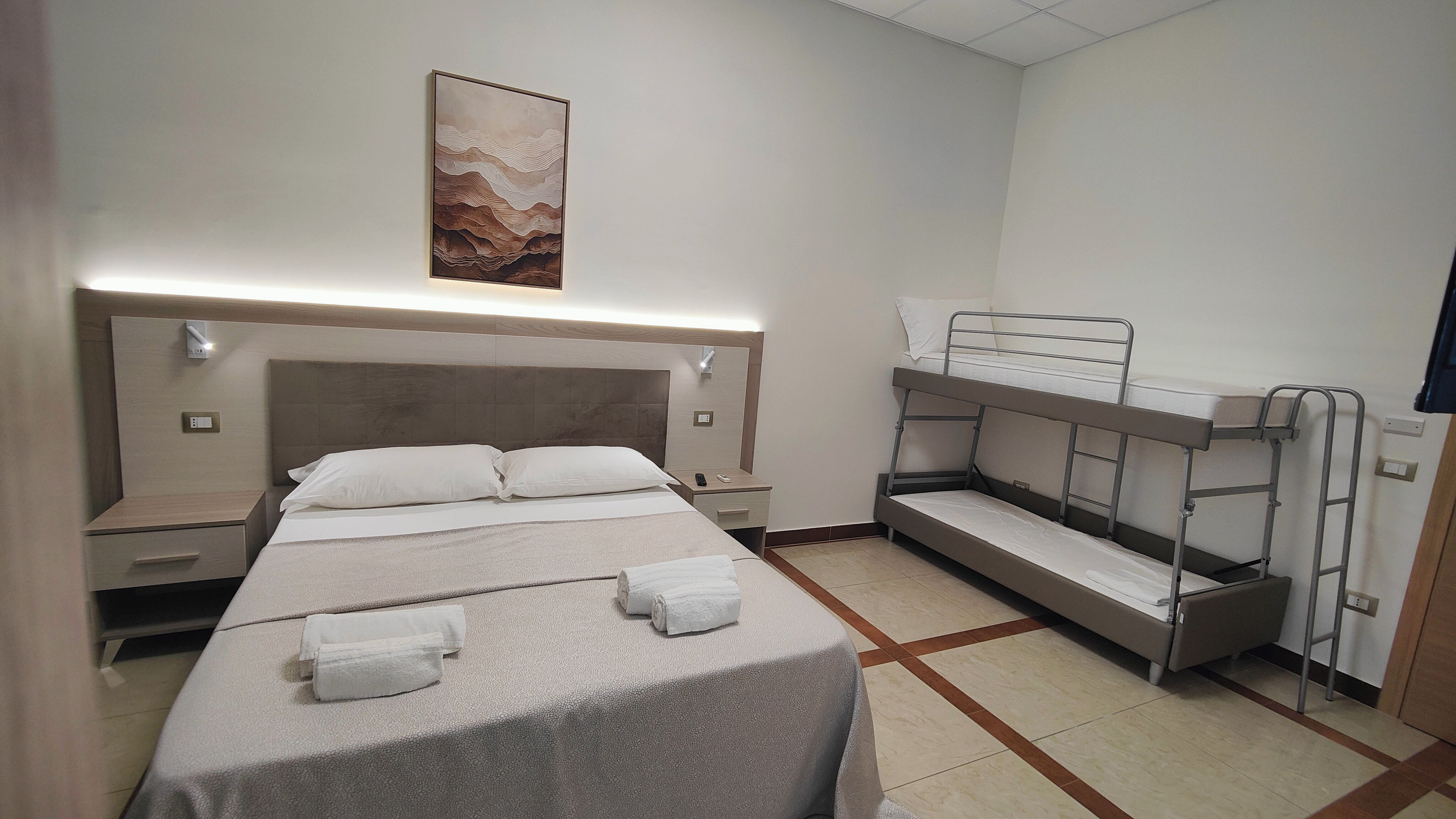 A hotel room with a bunk bed, a single bed, a bedside table, a nightstand, a wall-mounted light, and a painting.