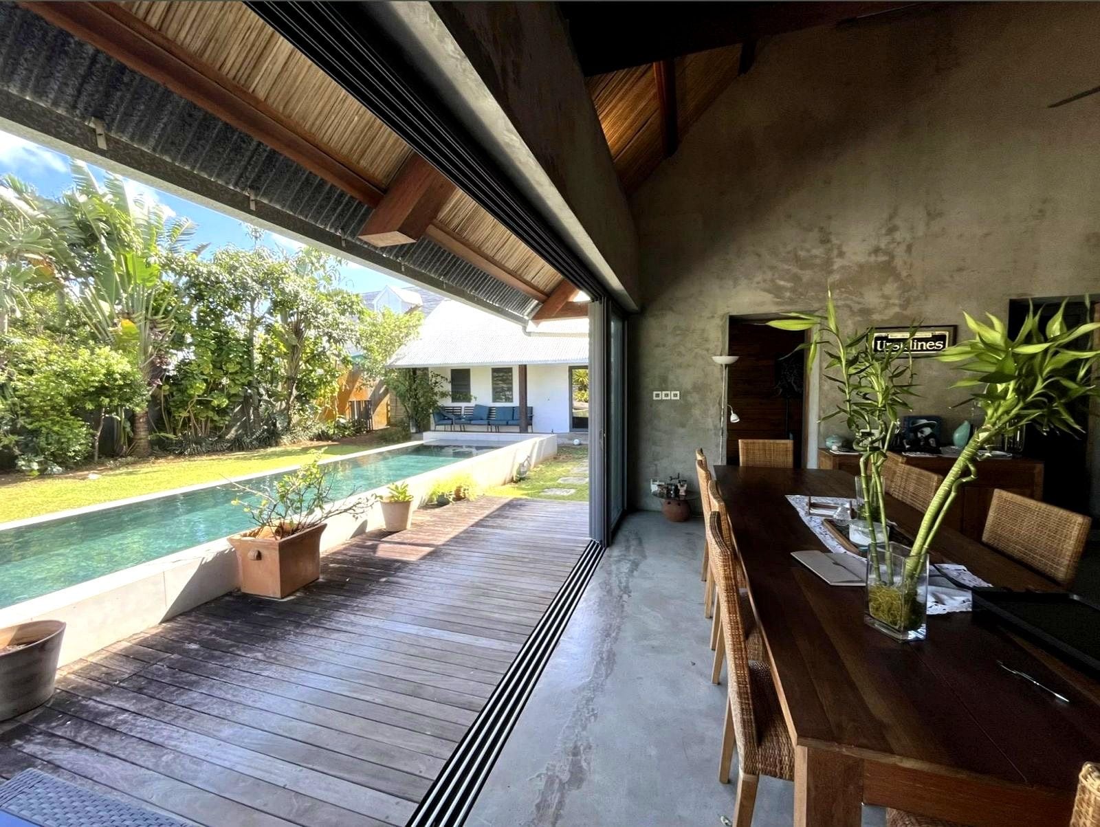 A modern indoor space with a pool, wooden deck, and a dining area with a view of the outdoors.