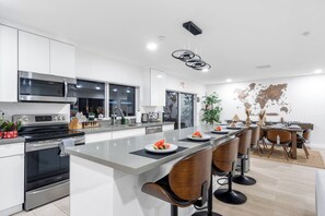 A modern kitchen with a central island, stainless steel appliances, and a dining area with wooden chairs. - Heated Pool + Backyard Fun + Sleeps 12 + 15 Min to the Beach (Hollywood)