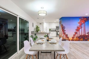Uma área de jantar moderna com uma mesa grande posta para seis pessoas, rodeada por cadeiras brancas. Uma cozinha visível ao fundo apresenta armários brancos e eletrodomésticos de aço inoxidável.