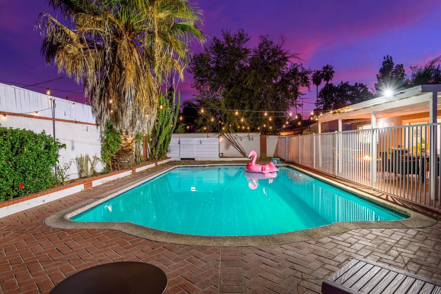 Piscina com boia em formato de flamingo rosa, cercada por um pátio de tijolos e uma cerca branca.