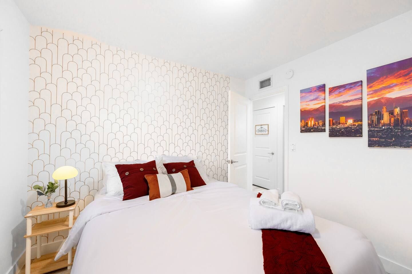 A hotel room with a patterned wall, a bed with pillows, a bedside table with a lamp, and three cityscape paintings on the wall.