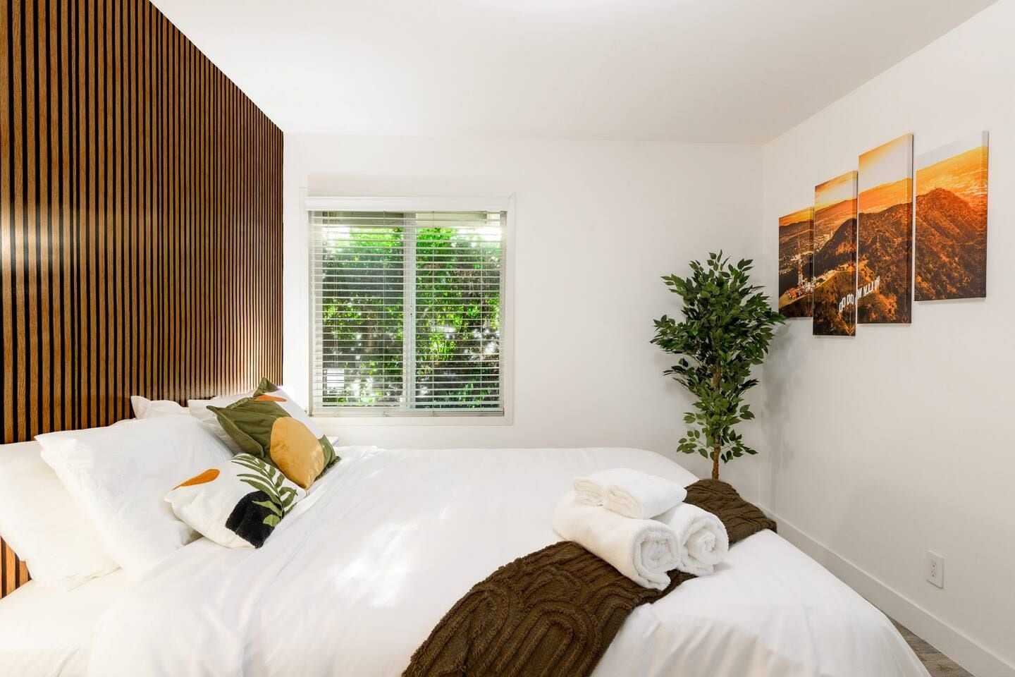 Quarto de hotel moderno com uma cama grande, janela com persianas, uma planta e um painel com três quadros de paisagens.