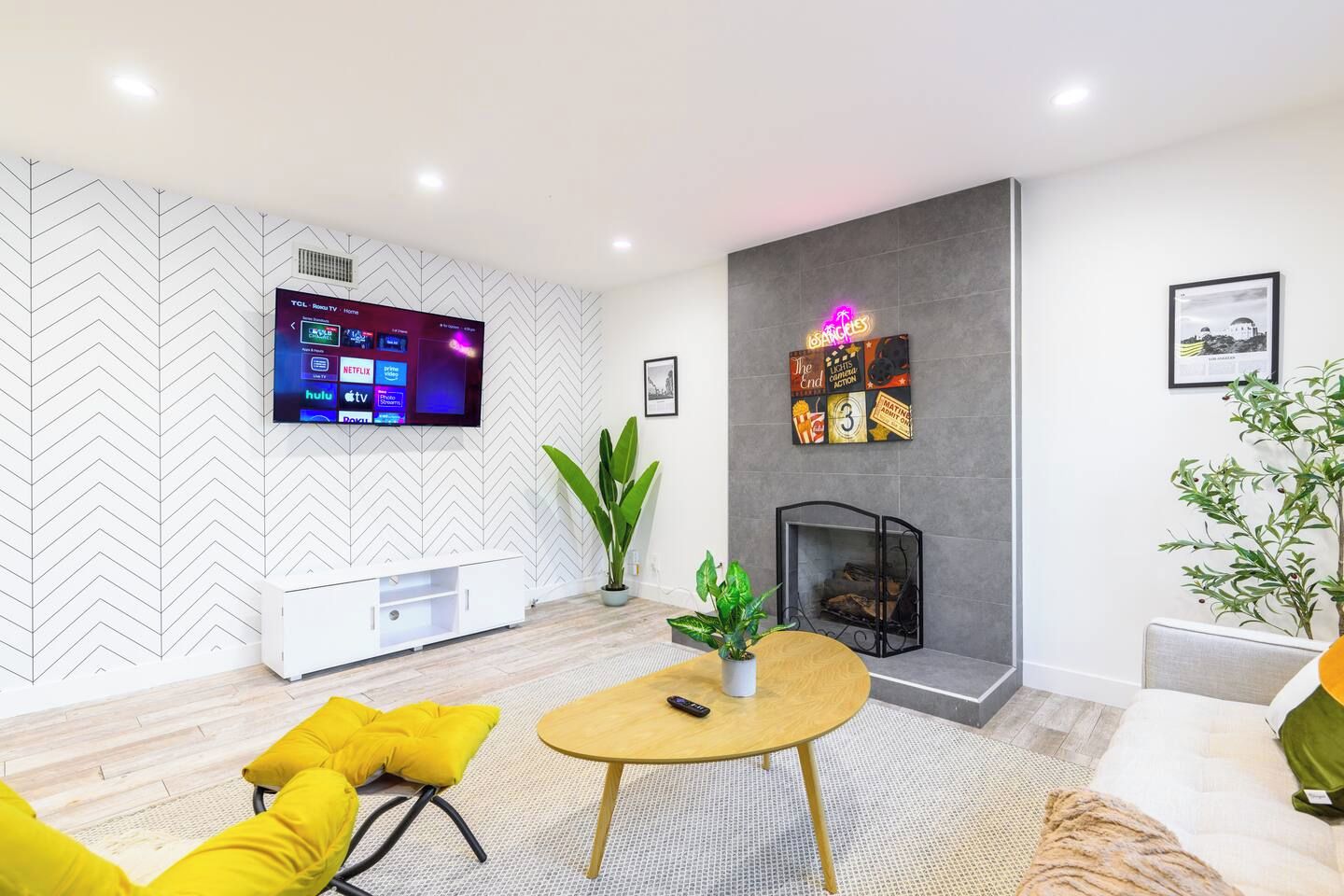 A modern living room with a flat-screen TV mounted on a white wall, a fireplace, a round coffee table, and yellow cushions.