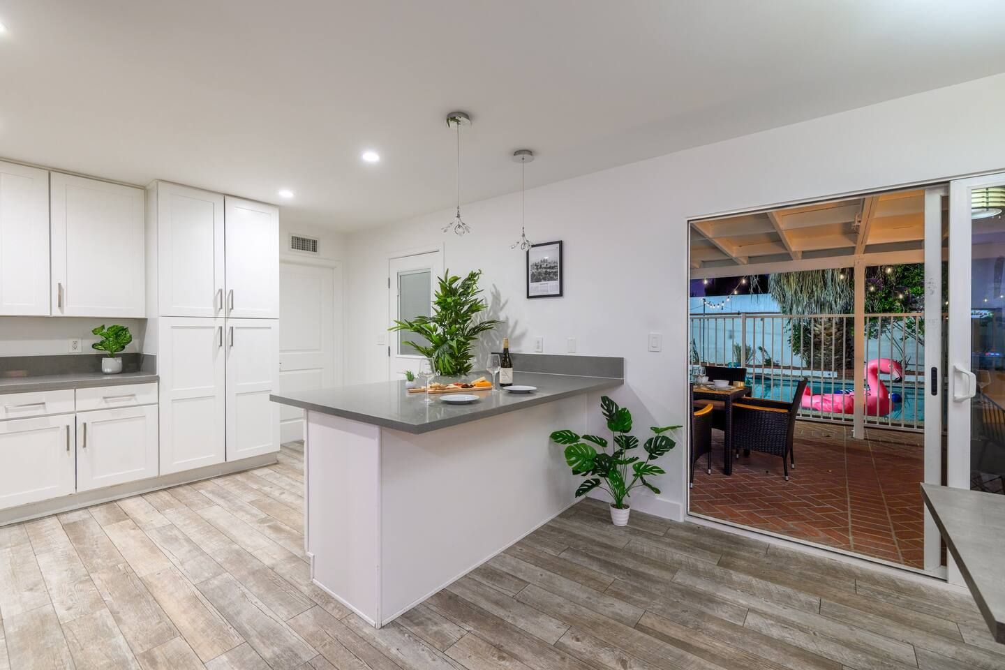 Cozinha moderna com armários brancos, bancada cinza e uma pequena ilha. Há uma área de jantar com mesa e cadeiras, e uma área de piscina visível através de uma porta aberta.