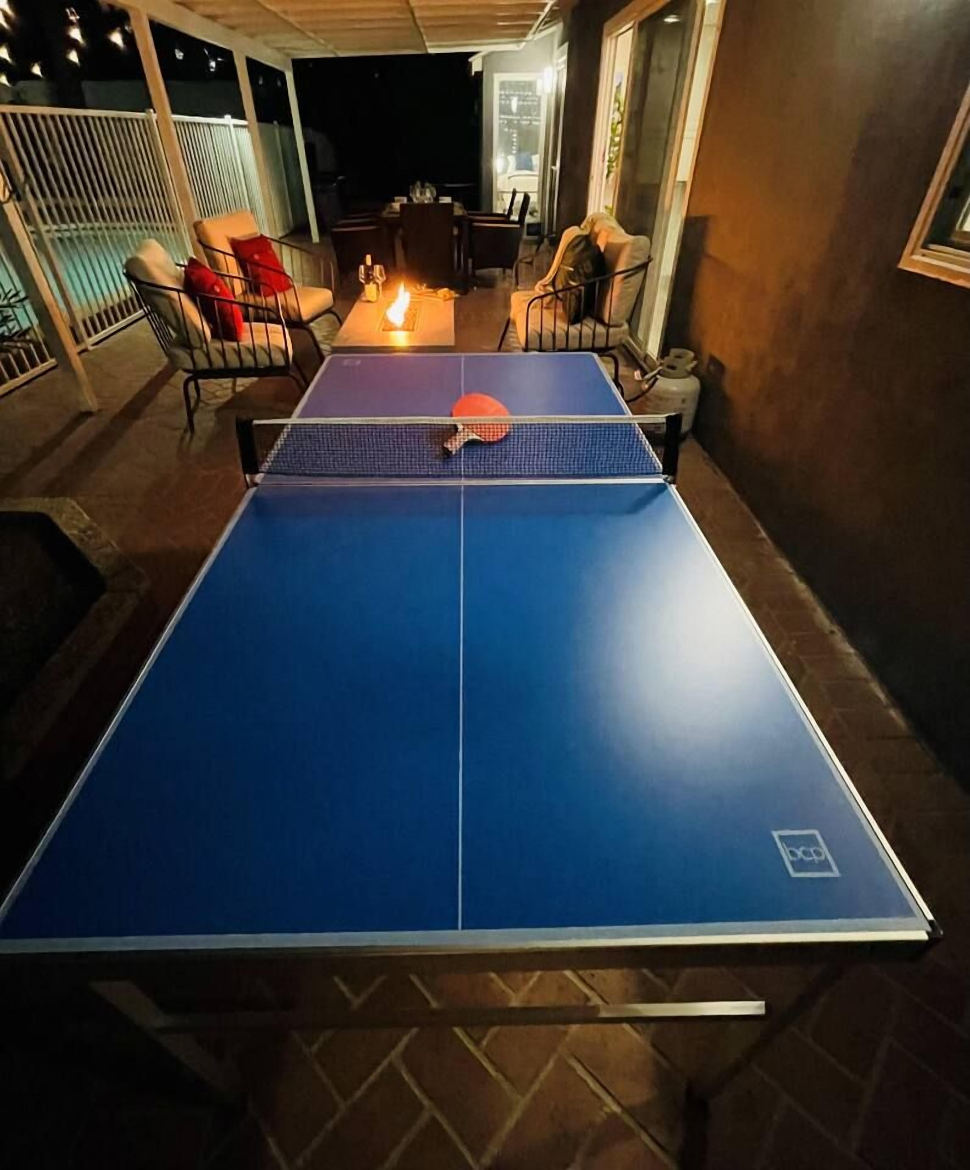Een tafeltennistafel met een blauw speeloppervlak en een net, omringd door stoelen en een tafel met een fles en een glas.