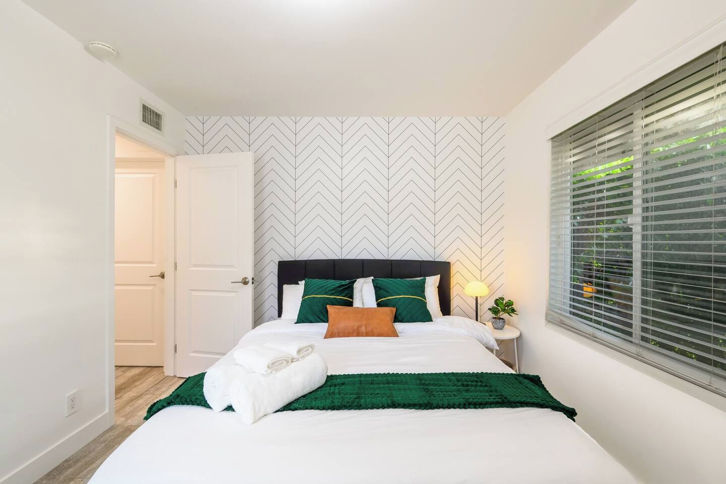 A neatly made bed with pillows and a blanket, a bedside lamp, and a window with blinds.