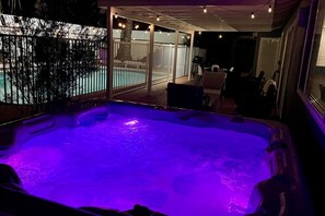 Uma banheira de hidromassagem com iluminação roxa, rodeada por um deck de madeira e uma porta de vidro que dá para uma área externa com cadeiras e uma mesa.