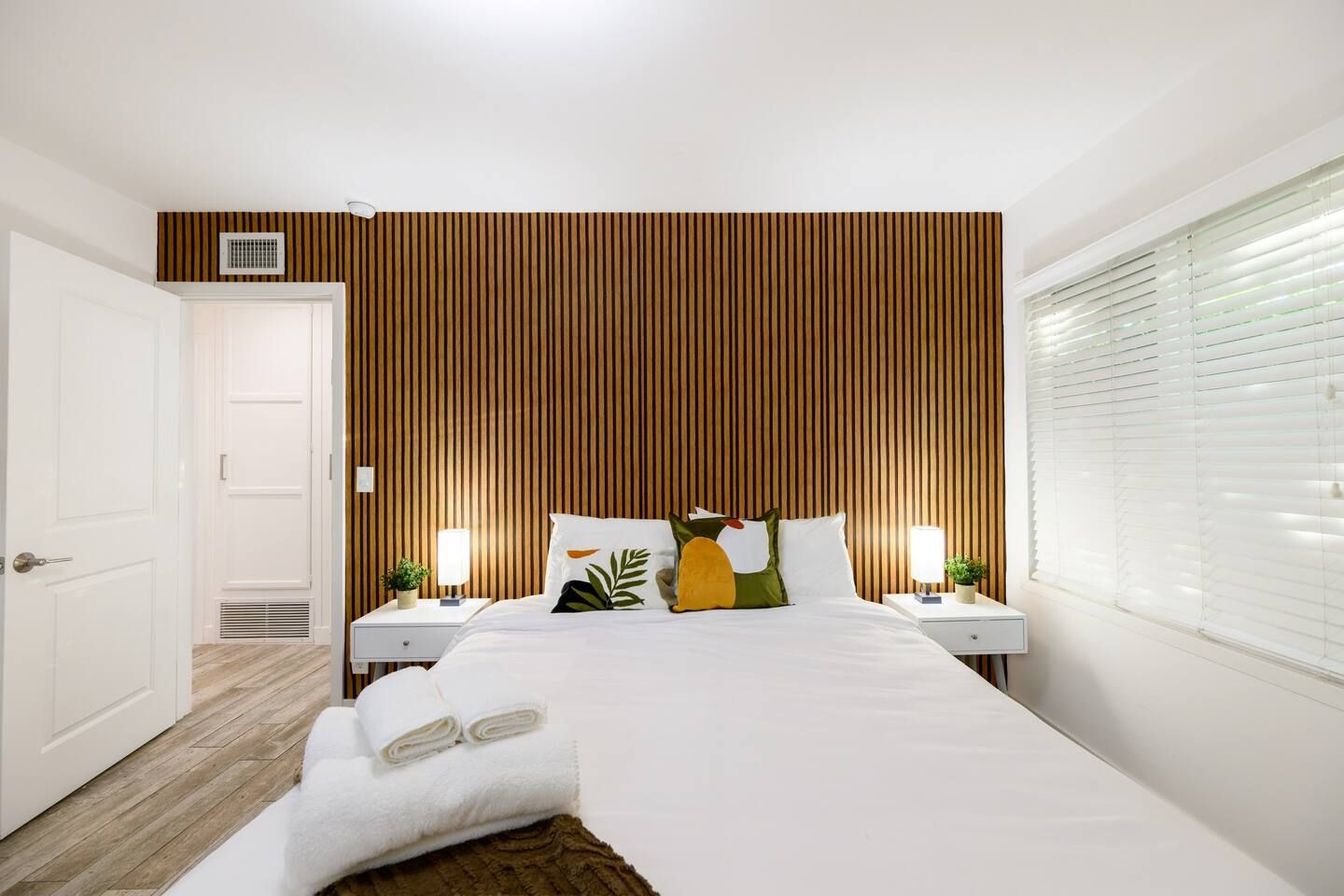 Een hotelkamer met een bed, nachtkastjes, een raam met zonwering, een deur en een wand met verticale houten latten.