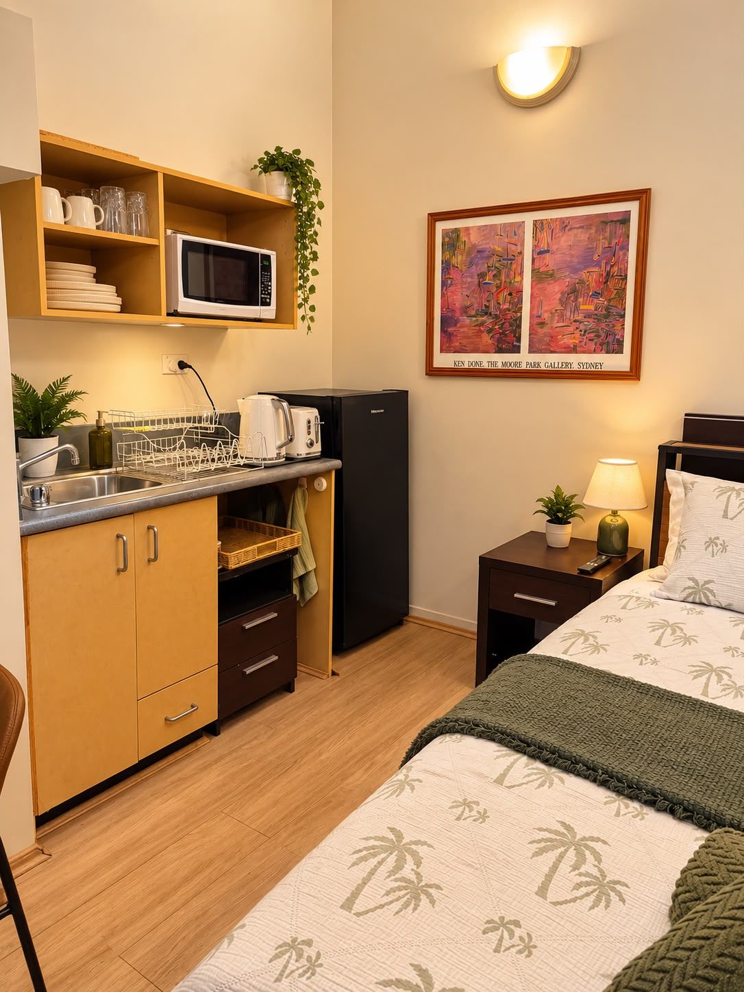 A compact hotel room with a kitchenette, a bed with a tropical-themed bedspread, a nightstand with a lamp, and a painting on the wall.