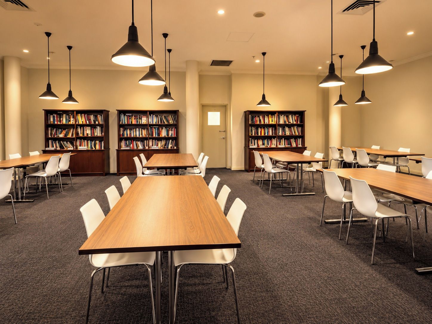 A conference room with multiple tables and chairs, overhead lighting, and bookshelves filled with books.