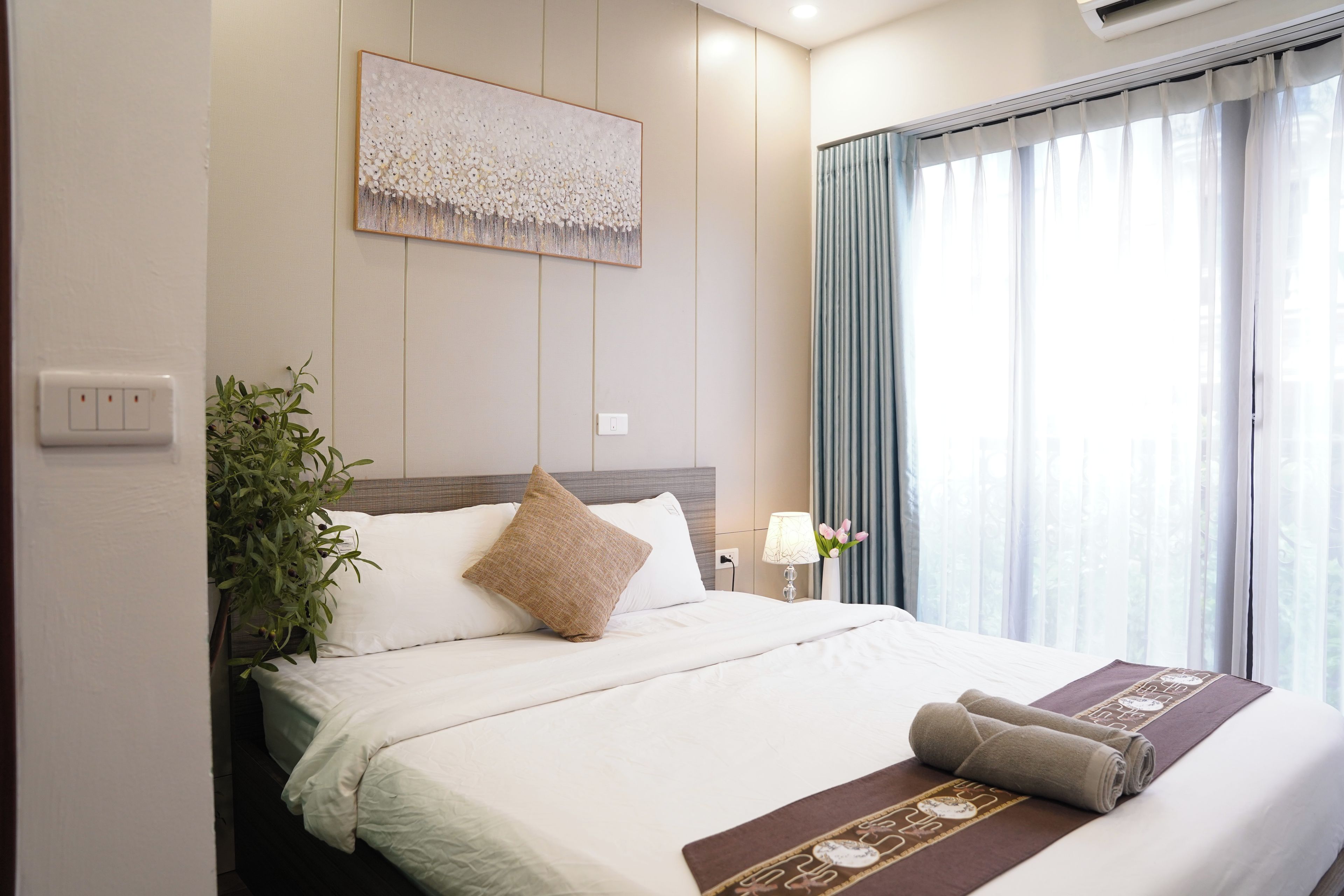 A hotel room with a bed, a wall-mounted artwork, a potted plant, a lamp, and a window with curtains.