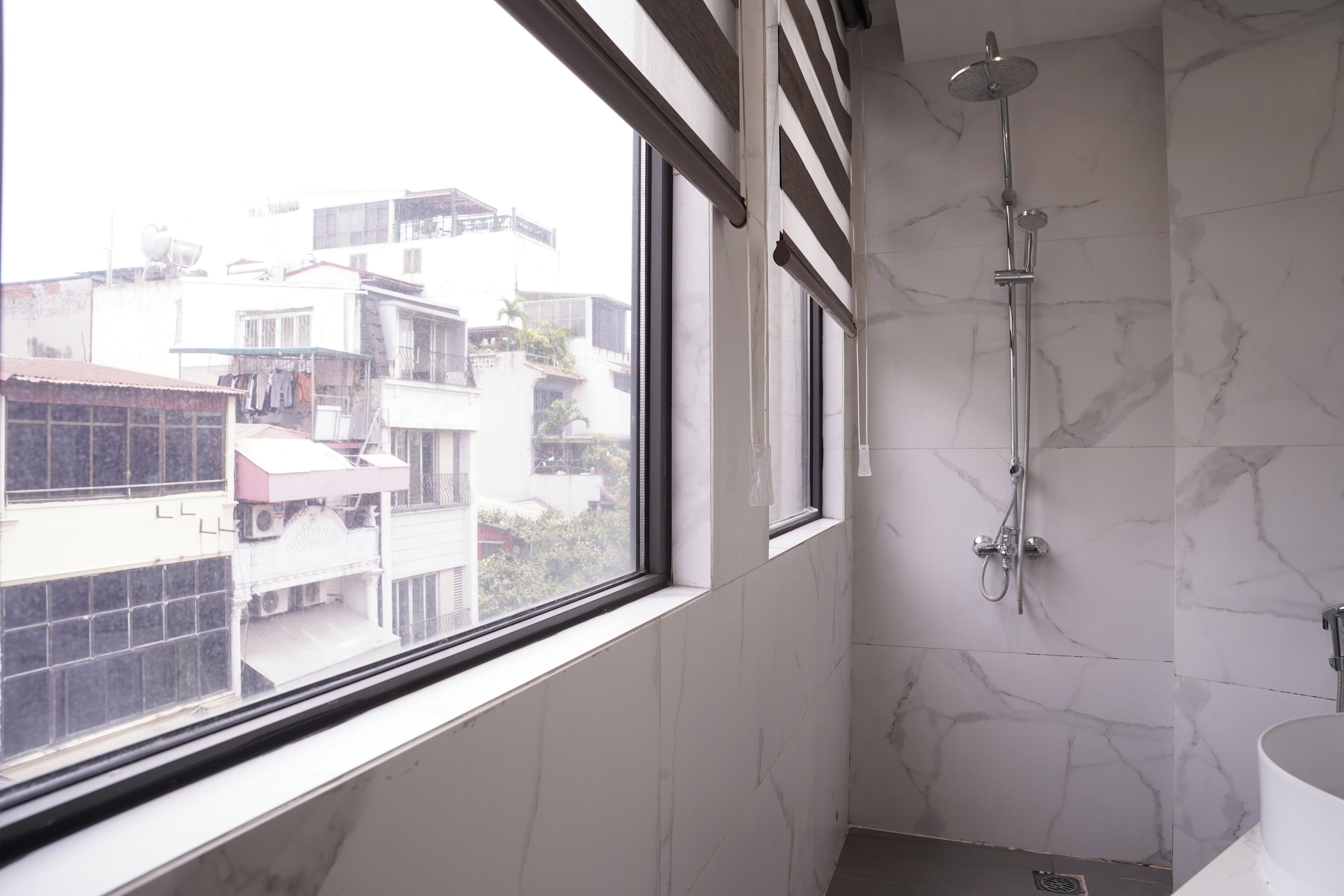 Casa de banho com chuveiro, pia e janela com vista para edifícios.