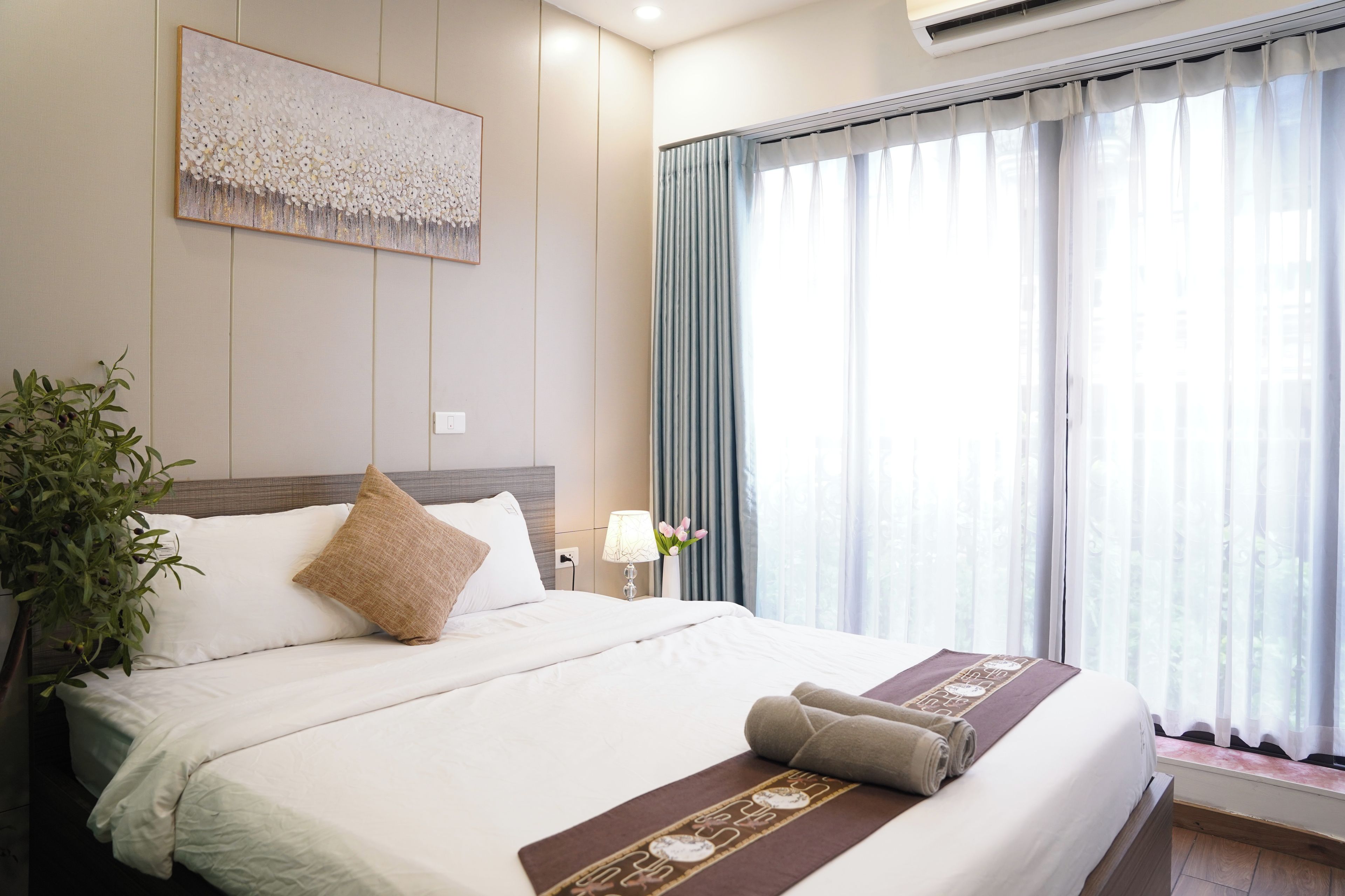 Ein Hotelzimmer mit Bett, Nachttisch, Lampe, Pflanze und einem fenster mit Vorhängen.