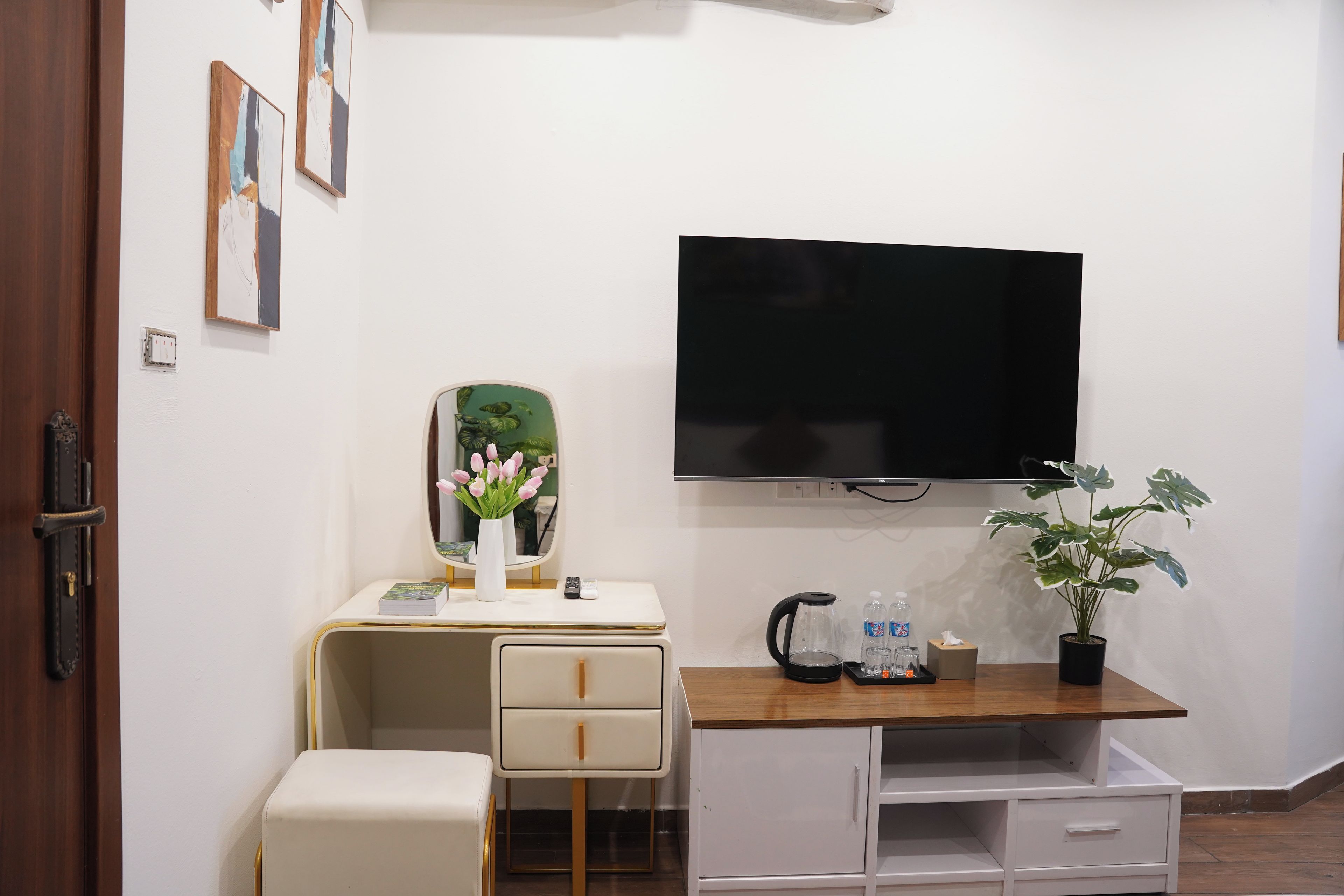 A modern room with a television mounted on the wall, a desk with a vase of flowers, a kettle, and a potted plant.